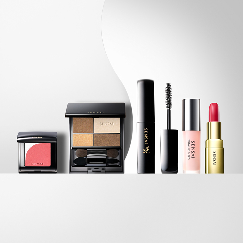 En rekke Sensai-sminkeprodukter er arrangert mot en hvit bakgrunn med en bølgende hvit detalj. Produktene omfatter en rouge, en øyenskyggepalett, en mascara, en lipgloss og en leppestift. Alle produktene har elegante forpakninger i svart, rosa og gull.