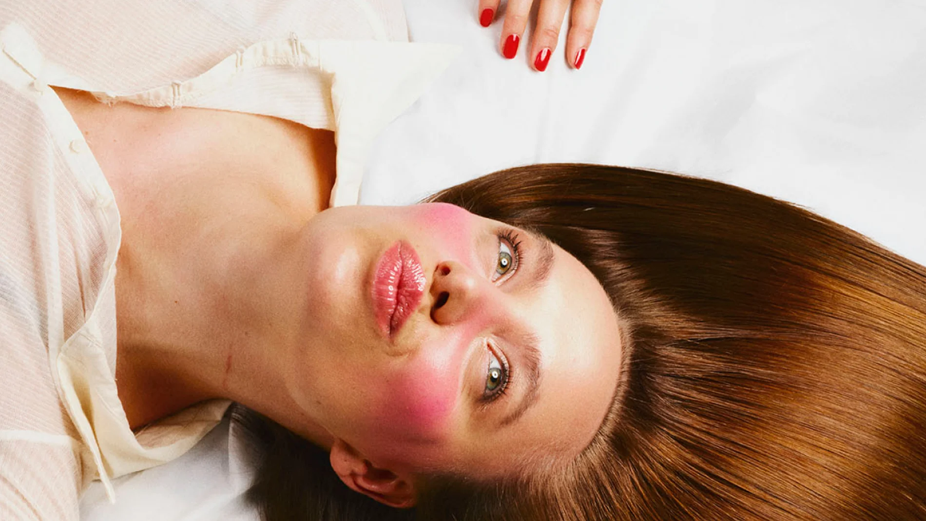 En person ligger ner mot vit bakgrund med långt, slätt brunt hår utspritt runt huvudet. Kindbenen är markerade med kraftigt rosa rouge och läpparna har en glansig, rosatonad finish. En hand med rödmålade naglar syns ovanför huvudet.