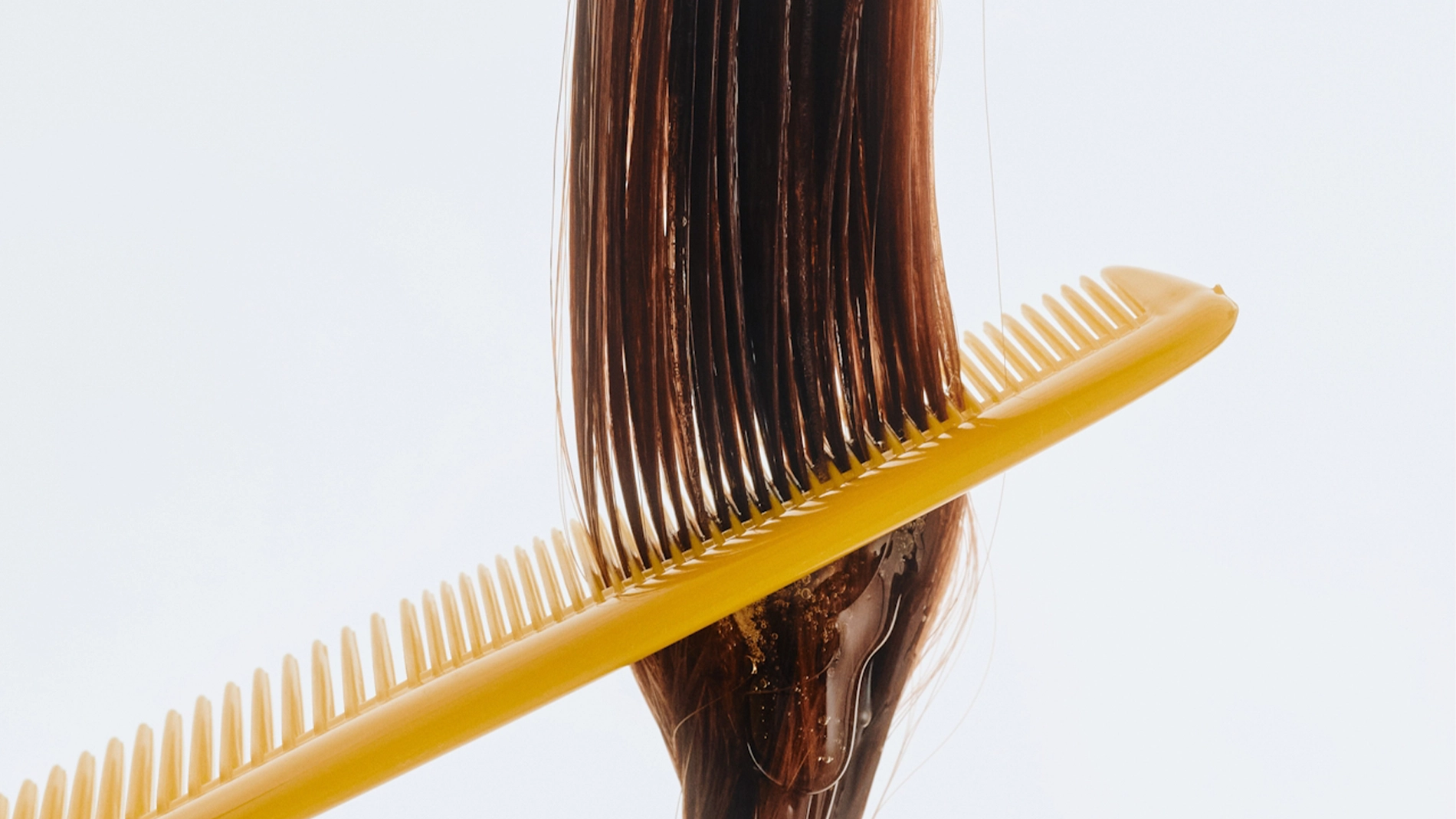 Lähikuva keltaisesta kammasta, joka vedetään pitkien, märkien ruskeiden hiusten läpi, ja hiusnaamio tai hoitoaine levittyy tasaisesti latvoihin. Painotus hiustenhoitoon ja hellävaraiseen selvitykseen vaaleaa taustaa vasten.