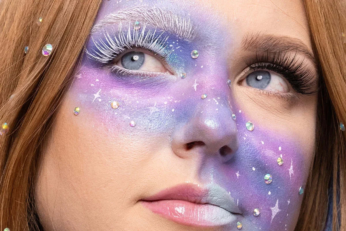Närbild av galaxinspirerad ansiktsmakeup i lila, blå och silver med vita fransar, skimrande stenar och målade stjärnor som skapar en drömlik effekt.