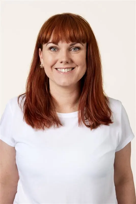 Portrett av en smilende person med rett rødt hår og hvit topp mot lys bakgrunn. Personen har naturlig makeup med markerte øyne og små øredobber.