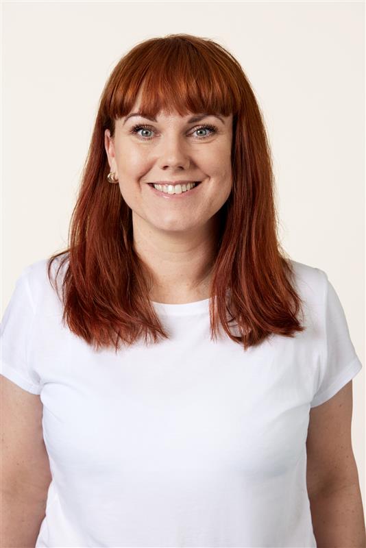 Porträtt av en leende person med rakt rött hår och vit topp mot ljus bakgrund. Personen bär naturlig makeup med markerade ögon och små örhängen.