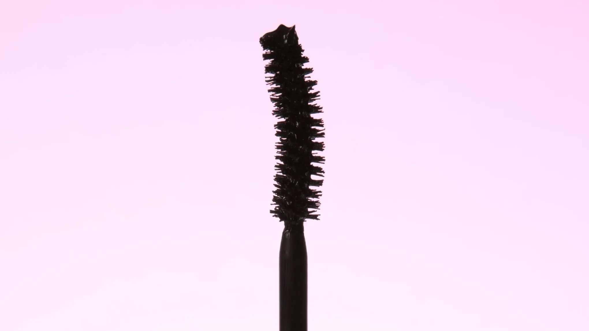 Et nærbilde av en mascarabørste med svart mascara mot en rosa bakgrunn.