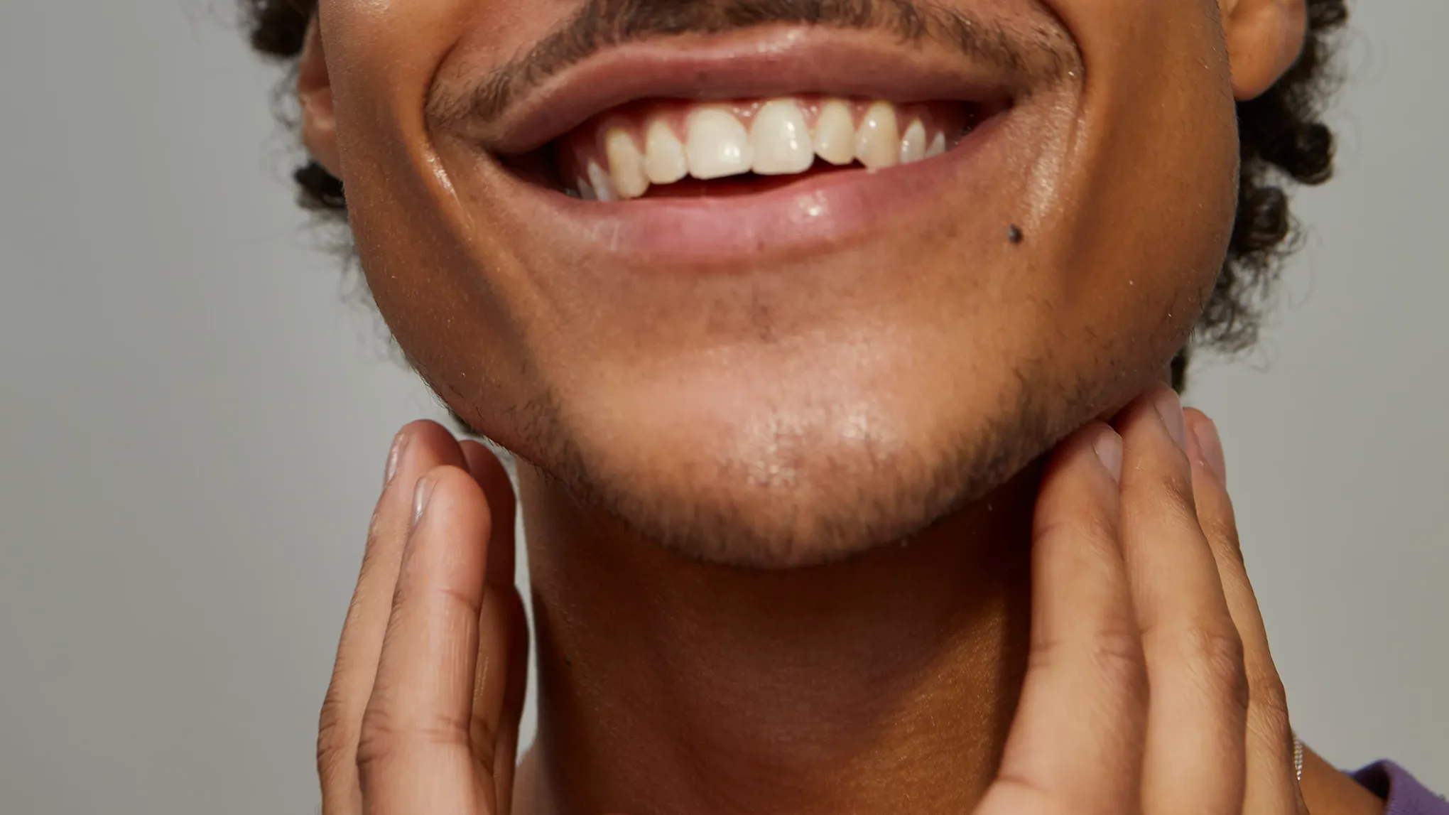 En man med mustasch som applicerar aftershave på halsen, mot en grågrön bakgrund.