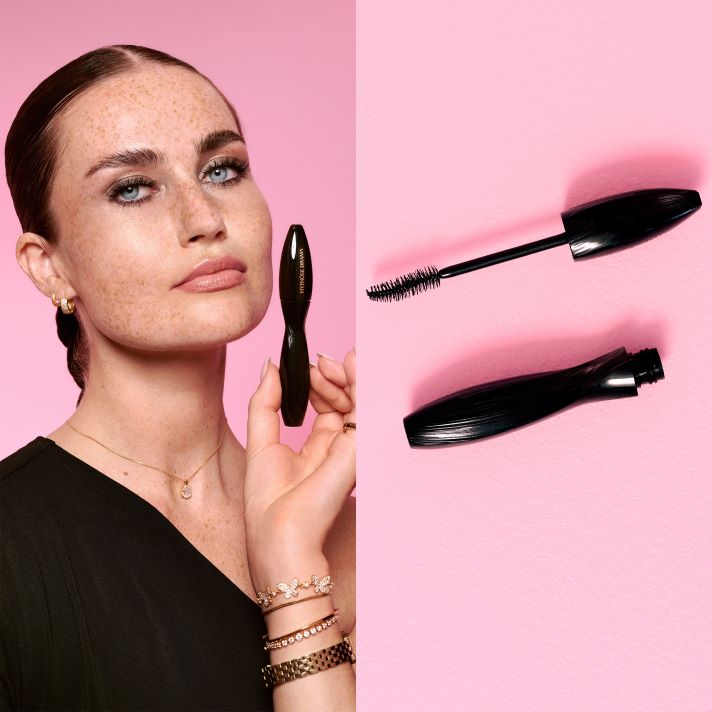 En person holder en Lancôme mascara foran ansiktet mot rosa bakgrunn, ved siden av vises mascarabørste og hylse separert.