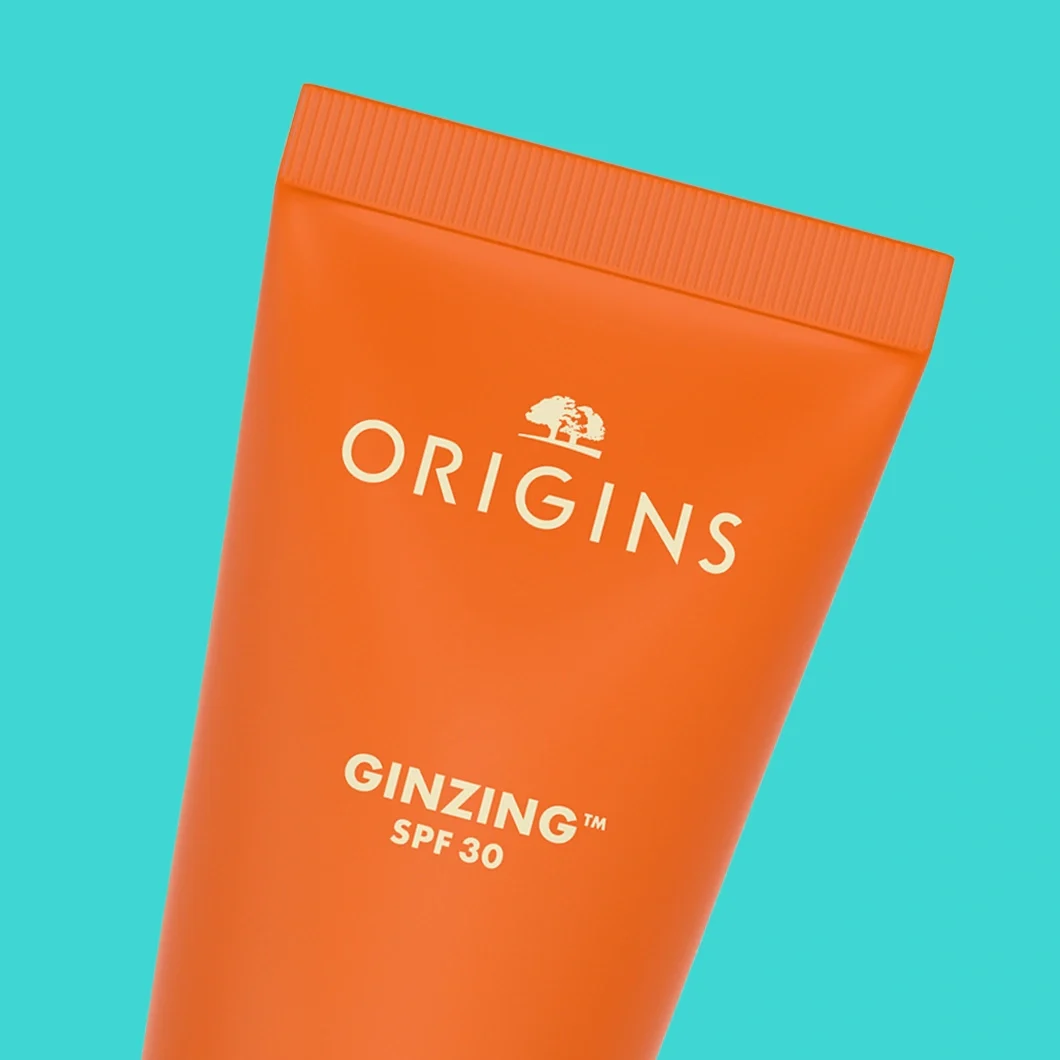 Origins GinZing SPF 30 on aurinkosuojallinen kasvovoide oranssissa tuubissa. Pakkauksessa näkyvät tuotemerkki "ORIGINS" ja teksti "GINZING™ SPF 30".