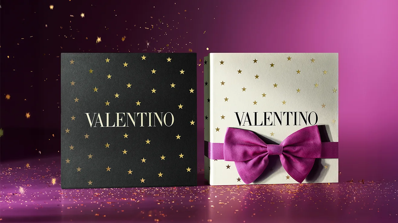 Kaksi Valentino couture-lahjapakkausta: valkoinen tähtikuvioinen Donna Born In Roma Intense ja musta maskuliininen Uomo Born In Roma EdT. Täydelliset joululahjat violetilla kimaltavalla taustalla.