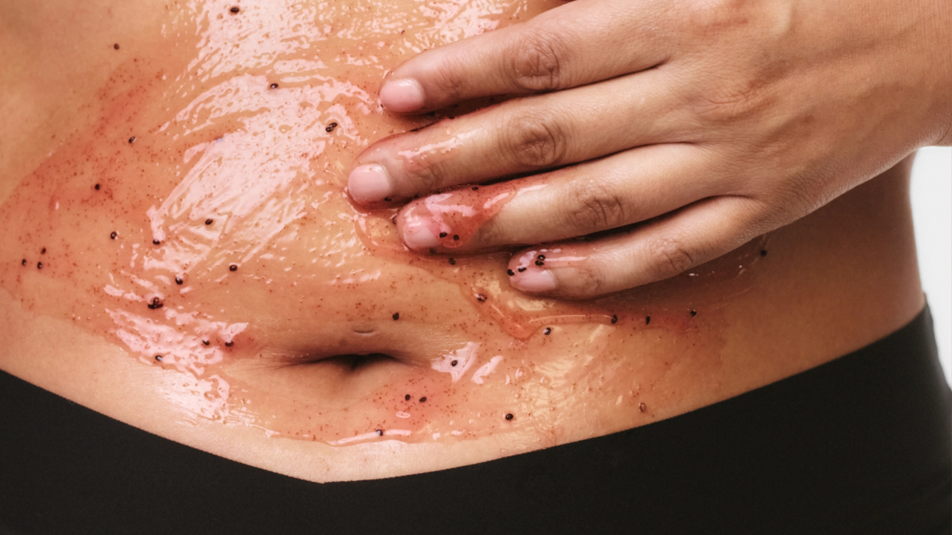 Lähikuva kädestä levittämässä vartalokuorintaa vatsan iholle, iholla kiiltävä pinta ja näkyviä kuorivia rakeita ihonhoidossa.