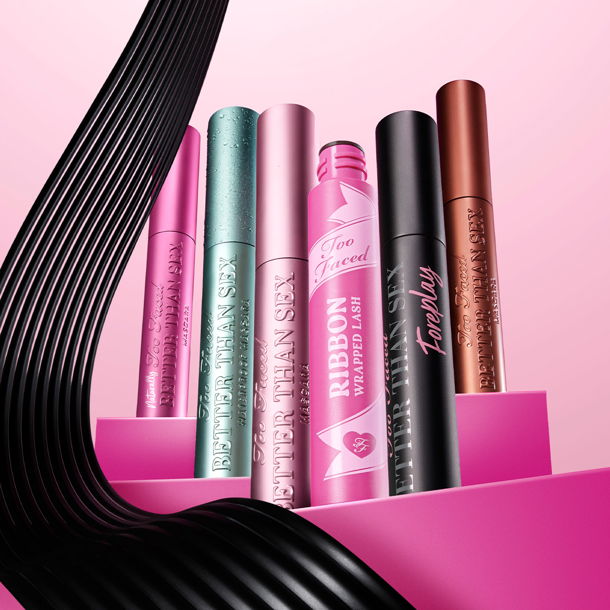Sex färgglada Too Faced-mascaror mot en rosa bakgrund. Från vänster till höger: rosa, grön, ljusrosa, rosa med bandmönster, svart och bronsfärgad.