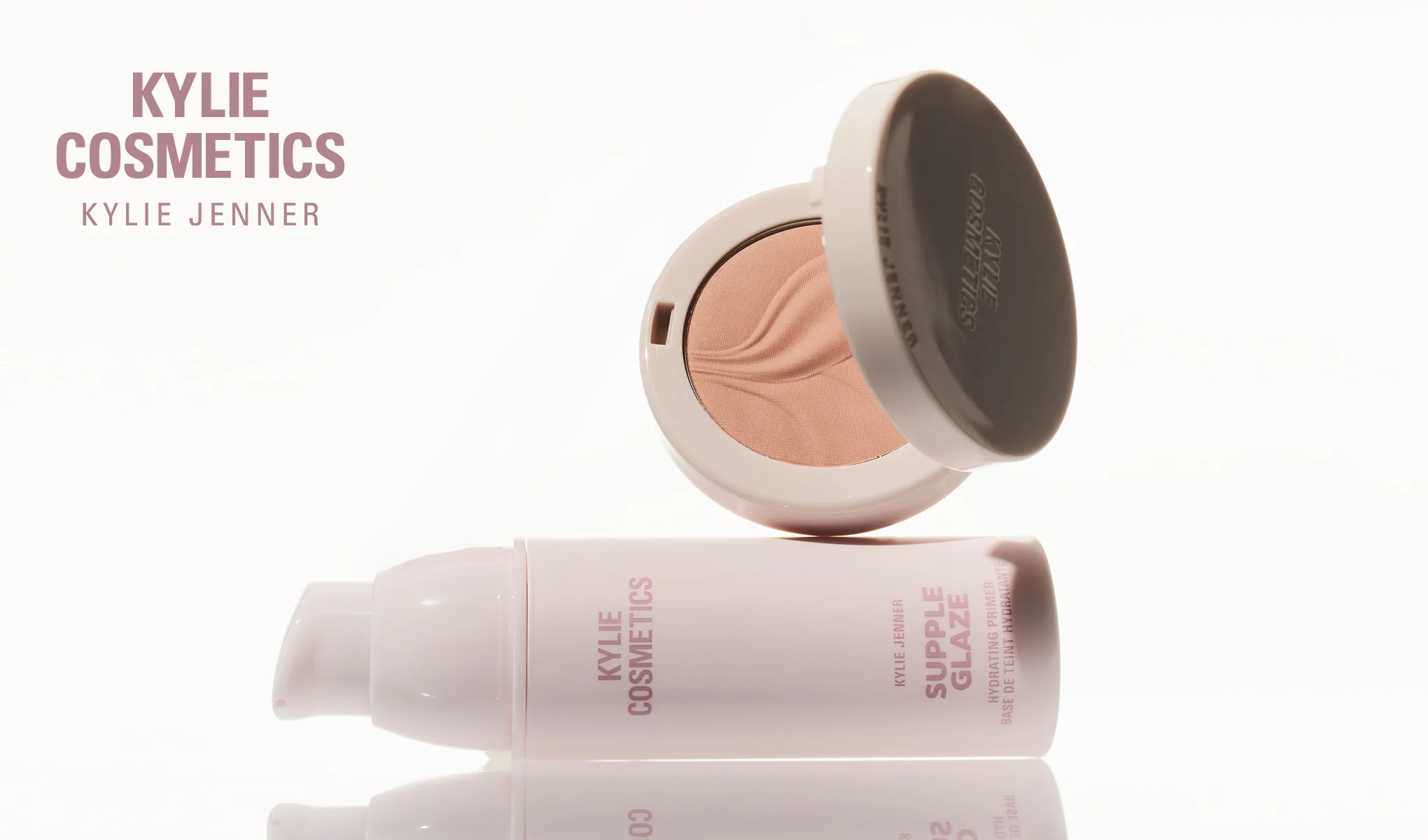 Kylie Cosmetics Supple Glaze Hydrating Primer og en åpen pudderkompakt vises mot en lys bakgrunn. Synlig tekst: "KYLIE COSMETICS", "KYLIE JENNER", "SUPPLE GLAZE", "HYDRATING PRIMER", "BASE DE TEINT HYDRATANTE" og "KYLIE COSMETICS" på produktene.