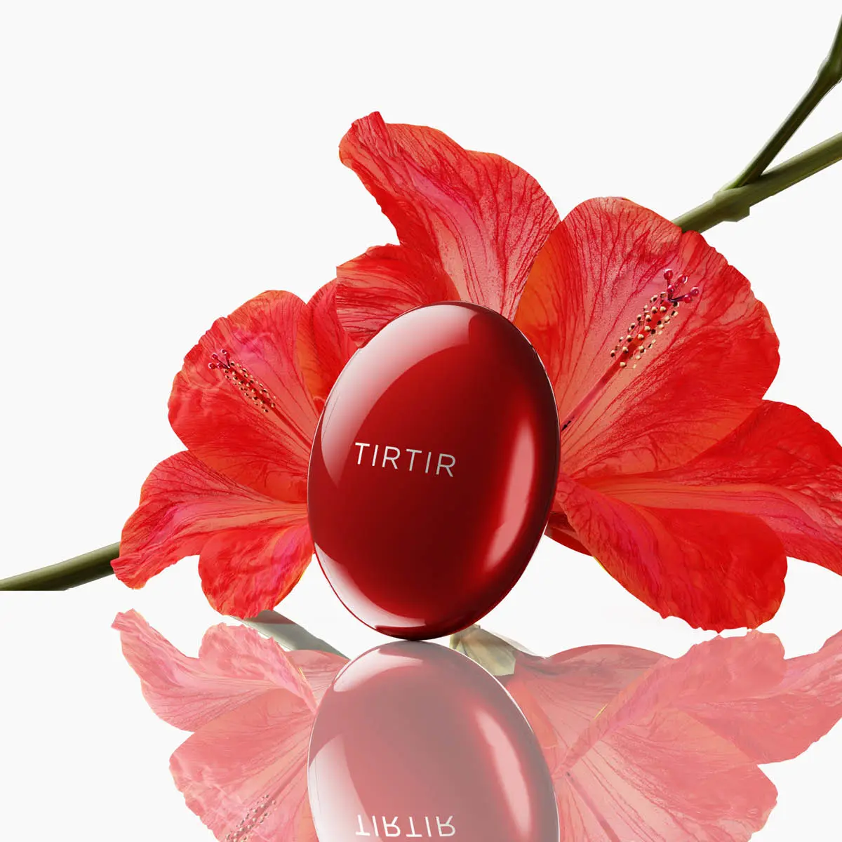 Rød TIRTIR cushion compact med merkenavn, omgitt av røde hibiskusblomster, reflektert på en overflate.
