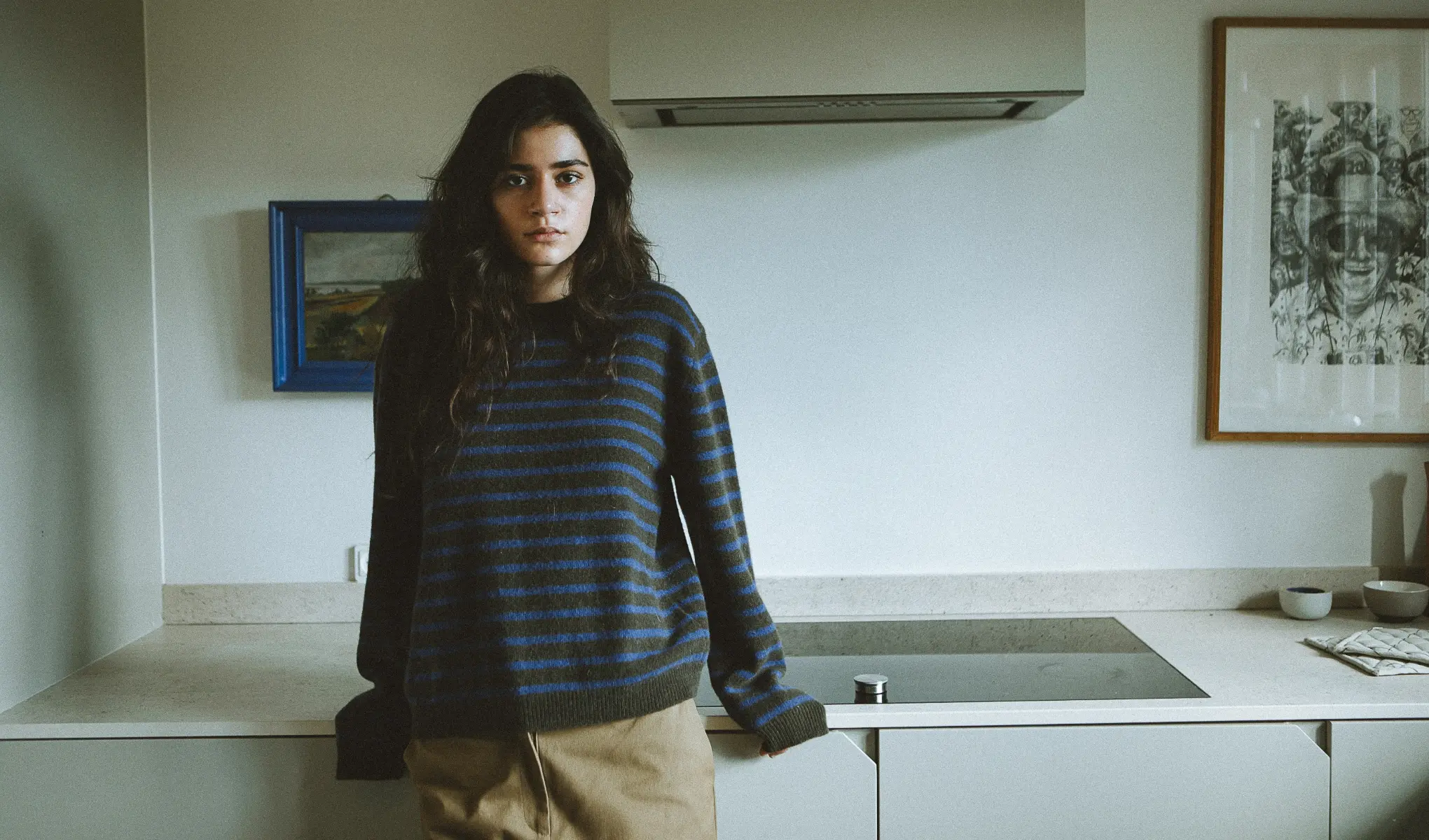 En person står i köket, iklädd en blå och svart randig tröja och beige byxor. I bakgrunden finns en spis, en målning med blå ram och en inramad svartvit teckning.