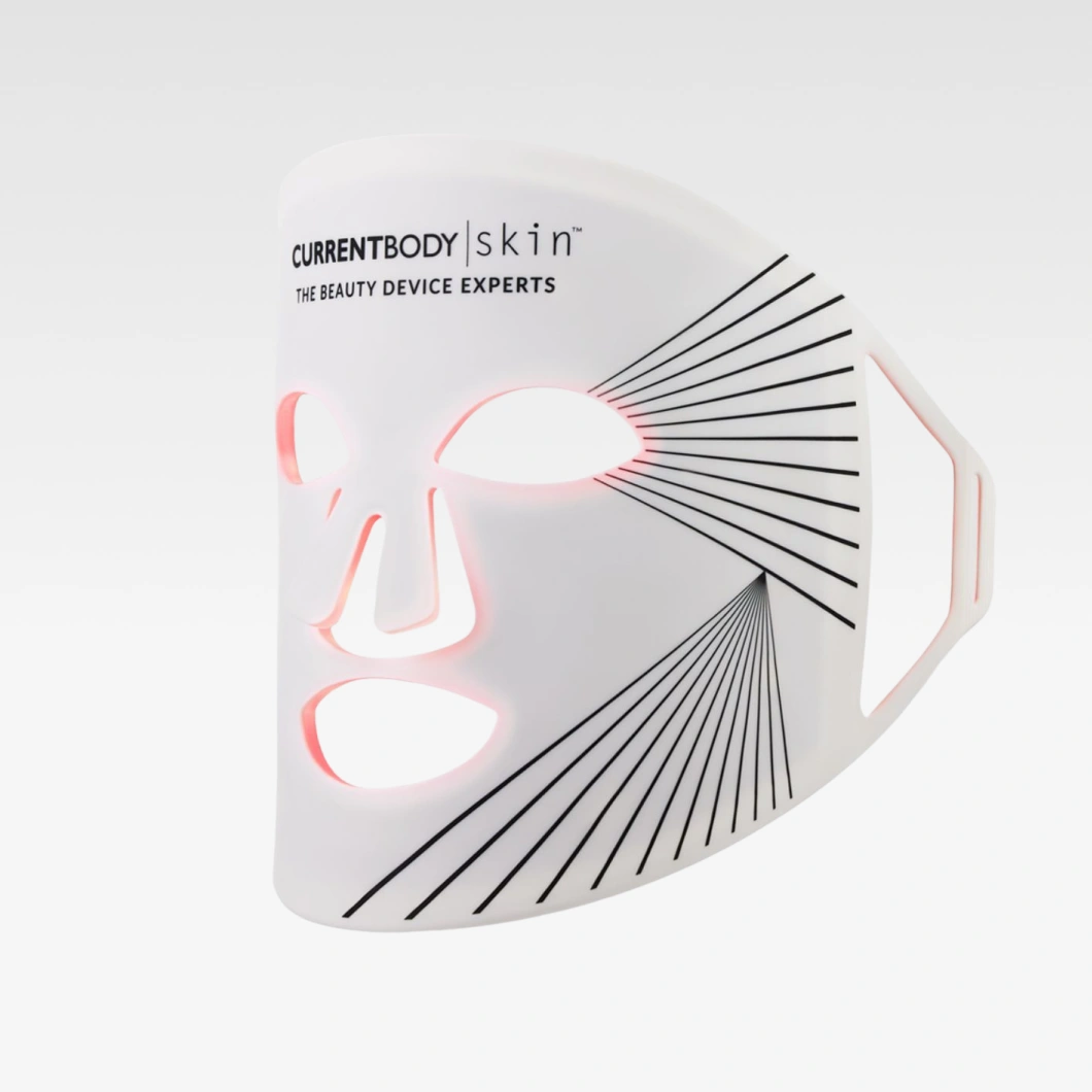 CurrentBody Skin LED-maske sammen med duftdiffuser, pillow spray, fotkrem, gua sha og wellnessprodukter mot lys blå bakgrunn. Produktene er arrangert i et spa-inspirert miljø med duftpinner, rosa sokker og beautyverktøy.