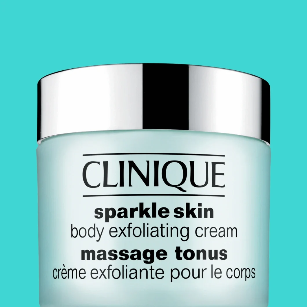 Clinique Sparkle Skin Body Exfoliating Cream -vartalokuorinta vaaleansinisessä purkissa hopeanvärisellä kannella turkoosia taustaa vasten. Pakkauksessa näkyvät tekstit "CLINIQUE", "sparkle skin", "body exfoliating cream", "massage tonus" ja "crème exfoliante pour le corps".
