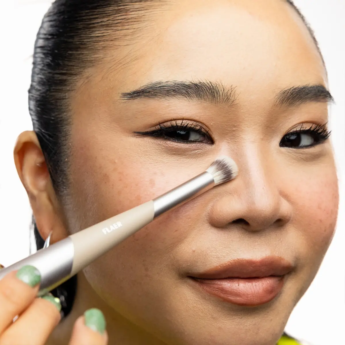 Nærbilde av person som påfører highlighter med en liten sminkekost på neseryggen. Sminkekosten har teksten FLAER på skaftet.