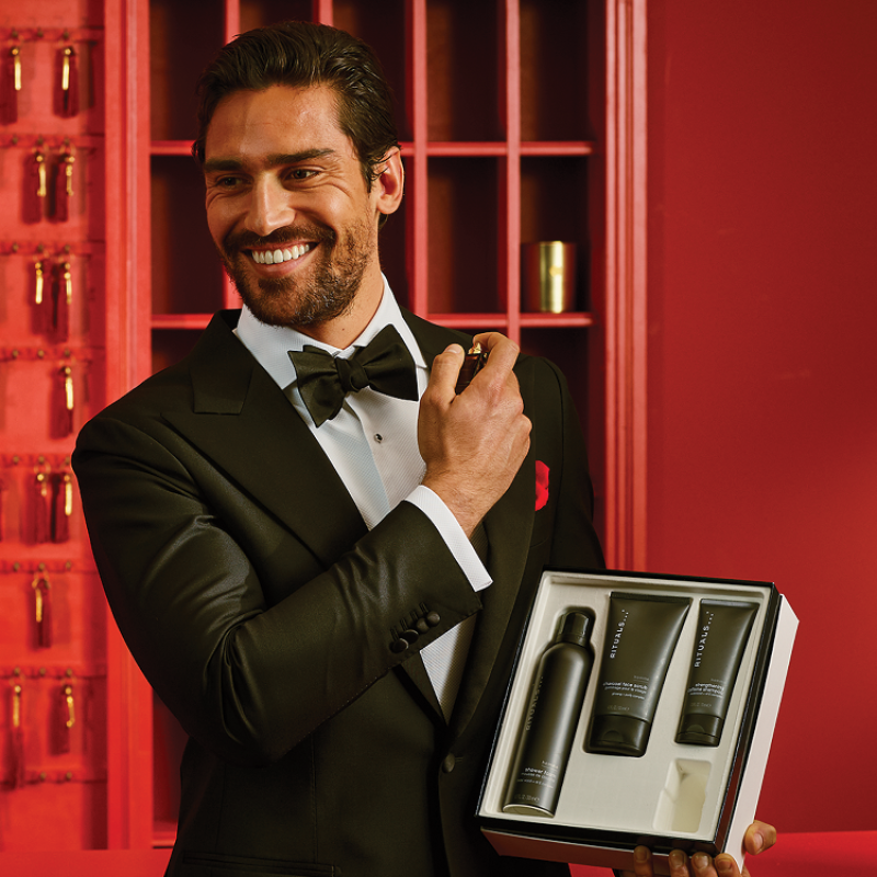 En man klädd i svart smoking och fluga som håller ett Rituals presentset. Presentsetet innehåller duschgel, kroppskräm och skrubbkräm i svarta förpackningar.