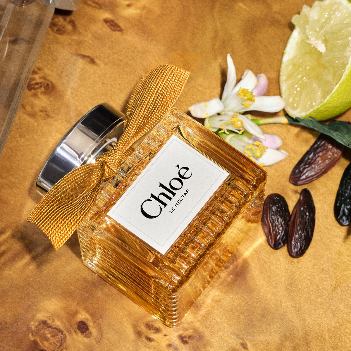Chloé Le Nectar -hajuvesi kultaisessa lasipullossa, jossa on kullanvärinen rusetti, leikatun limetin, valkoisten kukkien ja tummien siementen vieressä. Hajuvesipullossa näkyvät tekstit ”Chloé” ja ”LE NECTAR”.