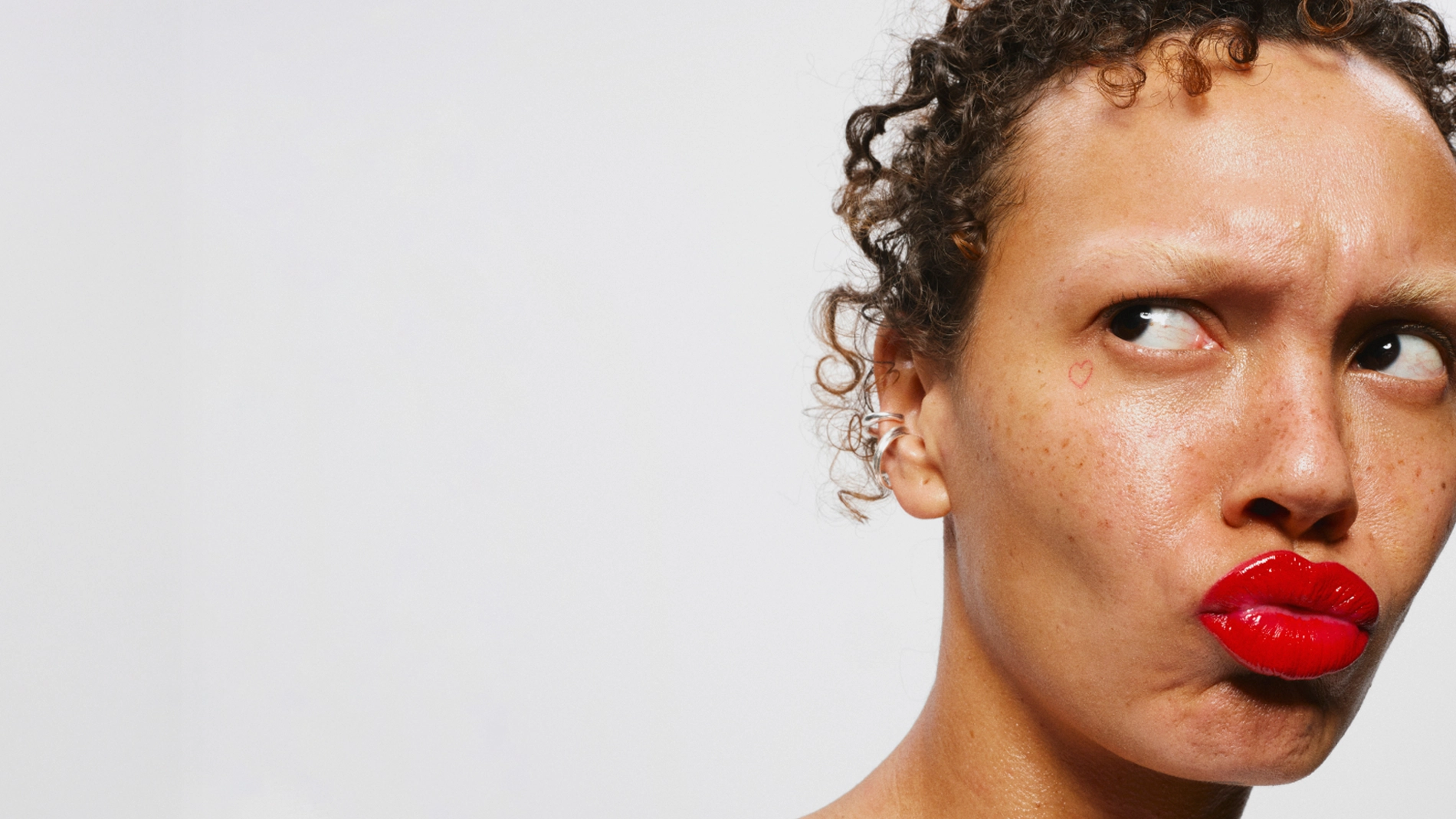 Henkilö, jolla on punaiset huulet ja luonnollinen meikki, katsoo sivulle otsa rypyssä; kasvoilla on pisamia ja pieni sydän poskella vaaleaa taustaa vasten.
