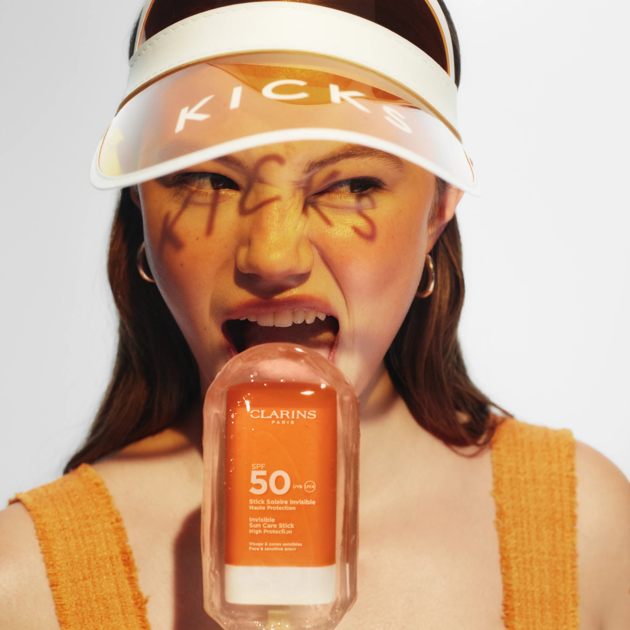 Person med solskjerm merket “KICKS” og tekst skrevet i ansiktet holder et solprodukt merket “CLARINS SPF 50 Invisible Sun Care Stick High Protection”.