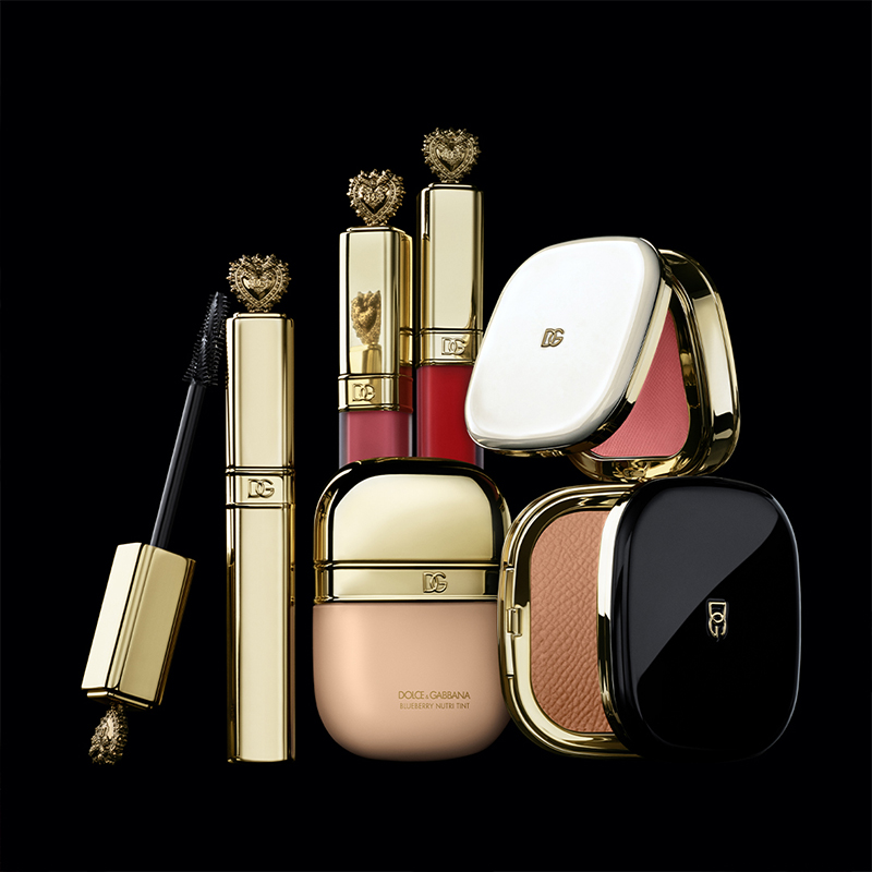Flera sminkprodukter från Dolce & Gabbana, placerade mot en svart bakgrund. Produkterna inkluderar mascara, läppstift, foundation och puder, alla med eleganta guldförpackningar och dekorativa detaljer. Läppstiften har hjärtformade toppar och puderdosorna har både vita och svarta lock med Dolce & Gabbana-logotypen. Produkterna är arrangerade på ett sätt som framhäver deras lyxiga design.