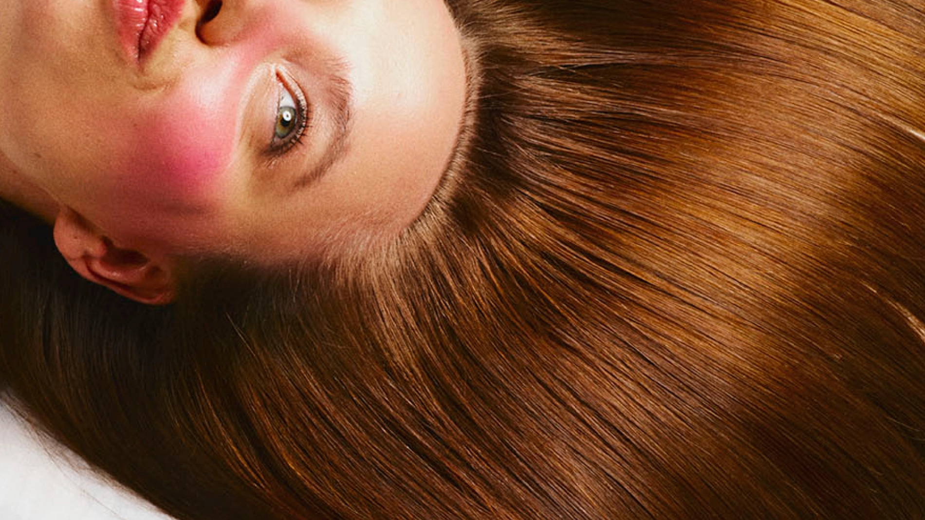 Närbild av en person som ligger ner med huvudet vänt åt sidan. Personen har långt, rakt brunt hår som breder ut sig runt huvudet. Ansiktet är synligt med rosatonad makeup på kinder och näsa samt glansiga läppar. Ljusa ögon och vit bakgrund.