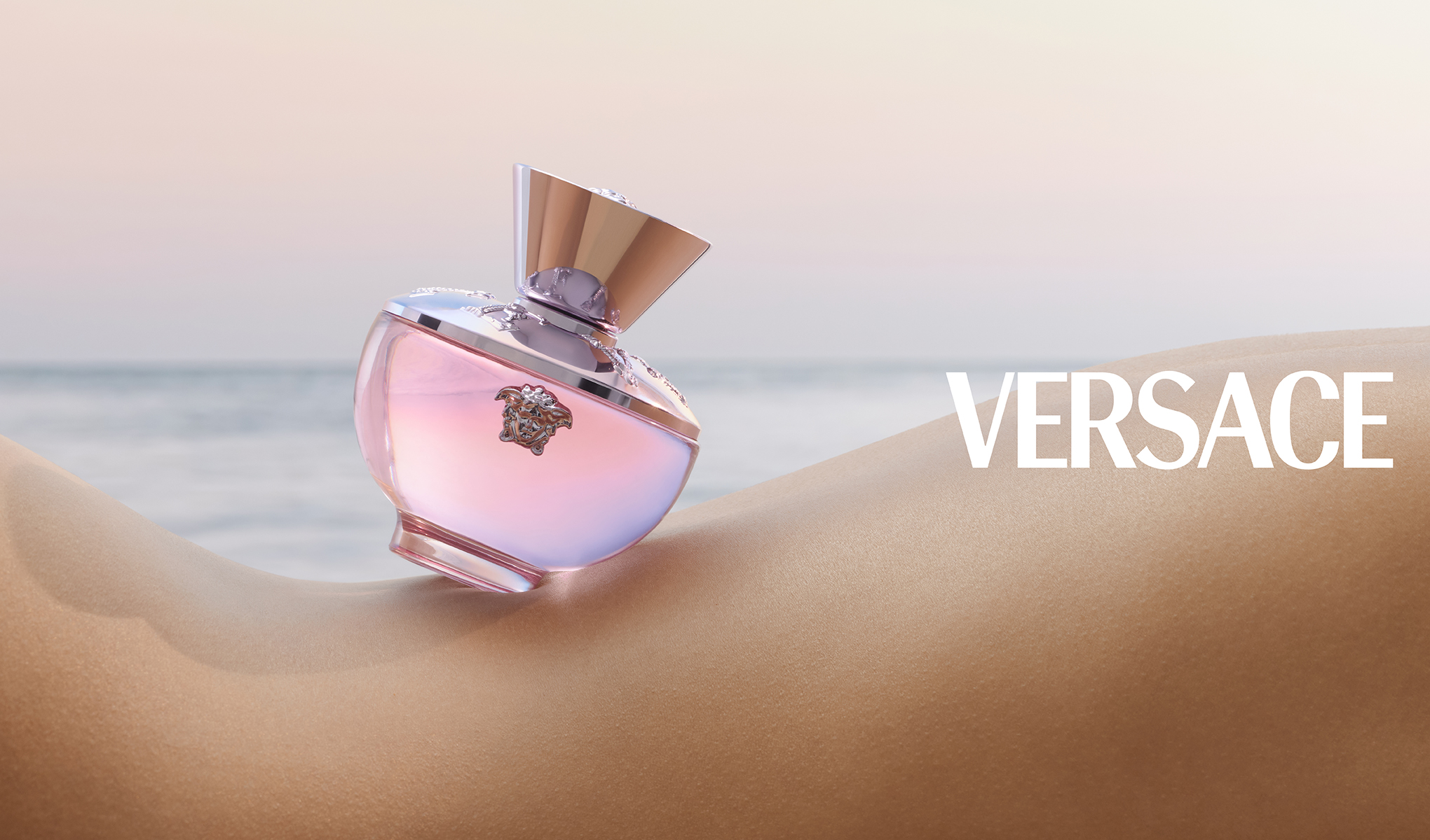 En rosa Versace-parfymeflaske hviler på menneskehud ved stranden. I bakgrunnen er det et rolig hav og himmel.