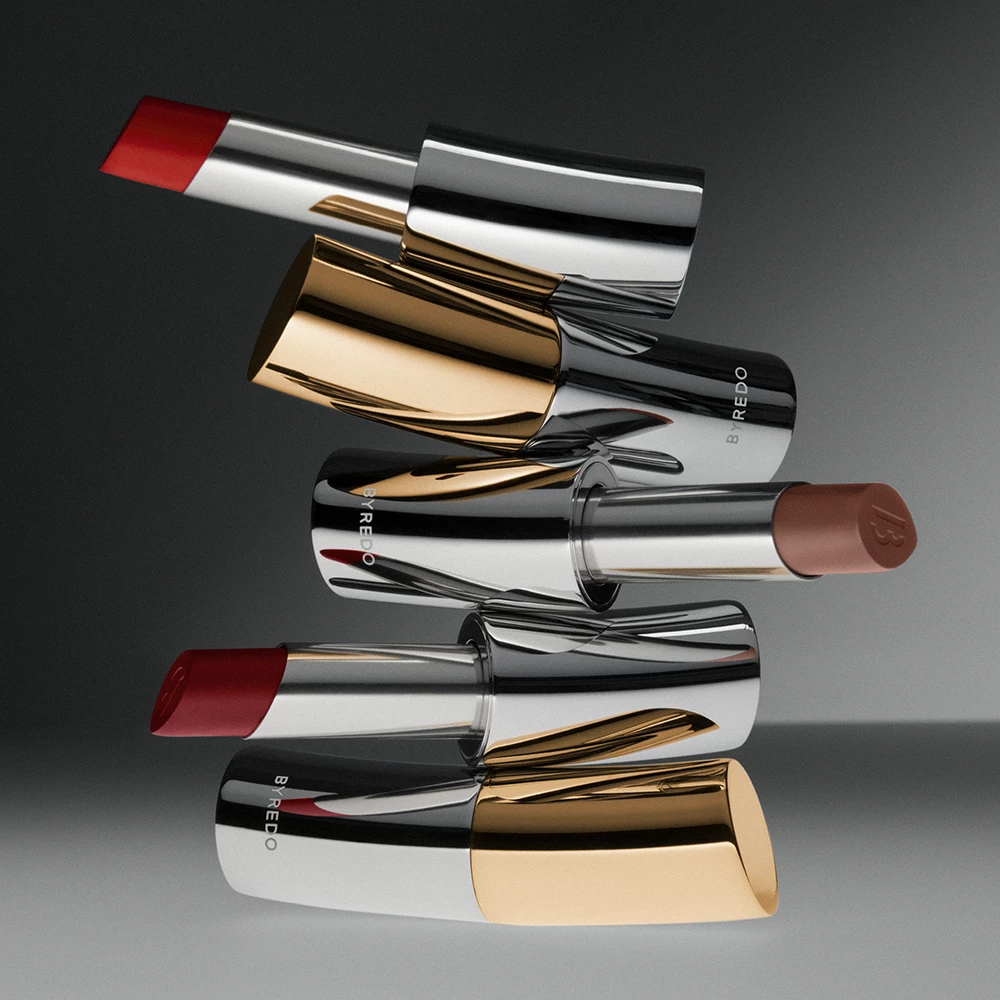 Fyra läppstift från Byredo, arrangerade i en stapel. Läppstiften har eleganta metallhylsor i silver och guld, och kommer i olika färger inklusive rött och brunt. Bakgrunden är grå.
