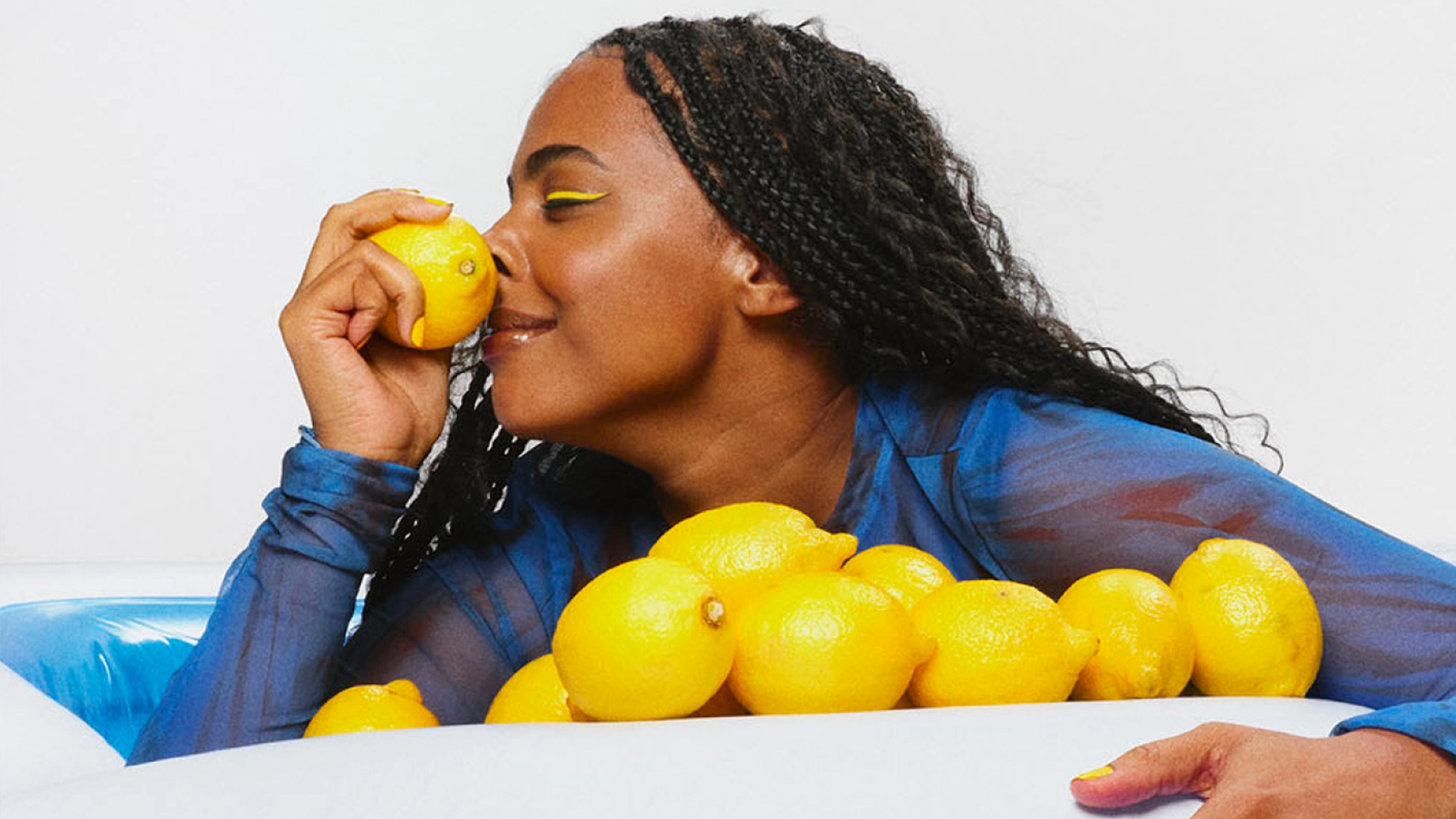En person med långt lockigt hår håller en citron mot näsan. Personen bär en genomskinlig blå långärmad topp och lutar sig mot ett vitt kar fyllt med flera gula citroner. Bakgrunden är ljus och enfärgad.
