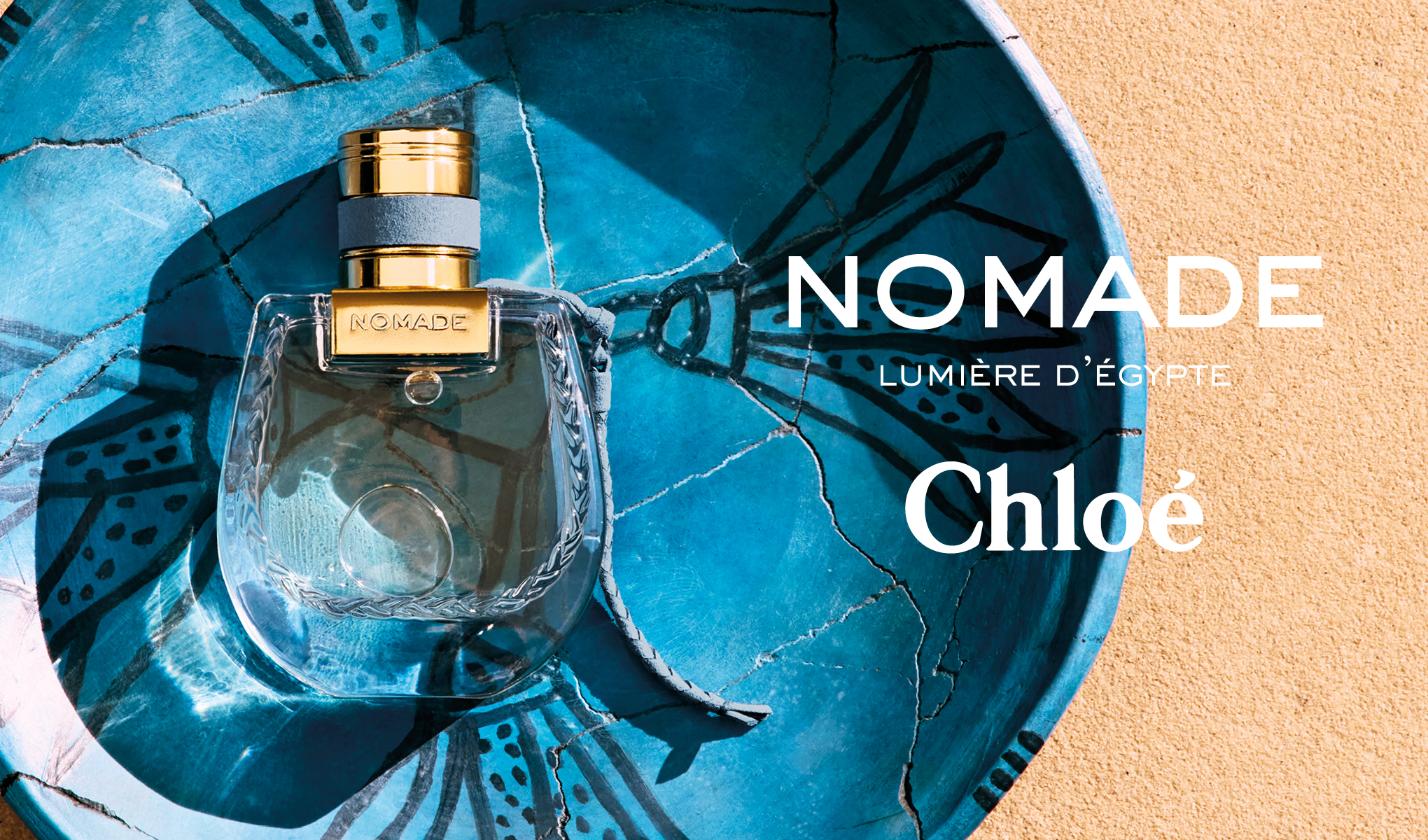 Chloén hajuvesipullo nimeltä ”Nomage Lumière D'égypte”, joka on sijoitettu siniseen kulhoon, jossa on koristeellinen kuvio. Taustalla on hiekanvärinen pinta. Teksti ”Nomade” ja ”Chloé” on sijoitettu pullon oikealle puolelle.