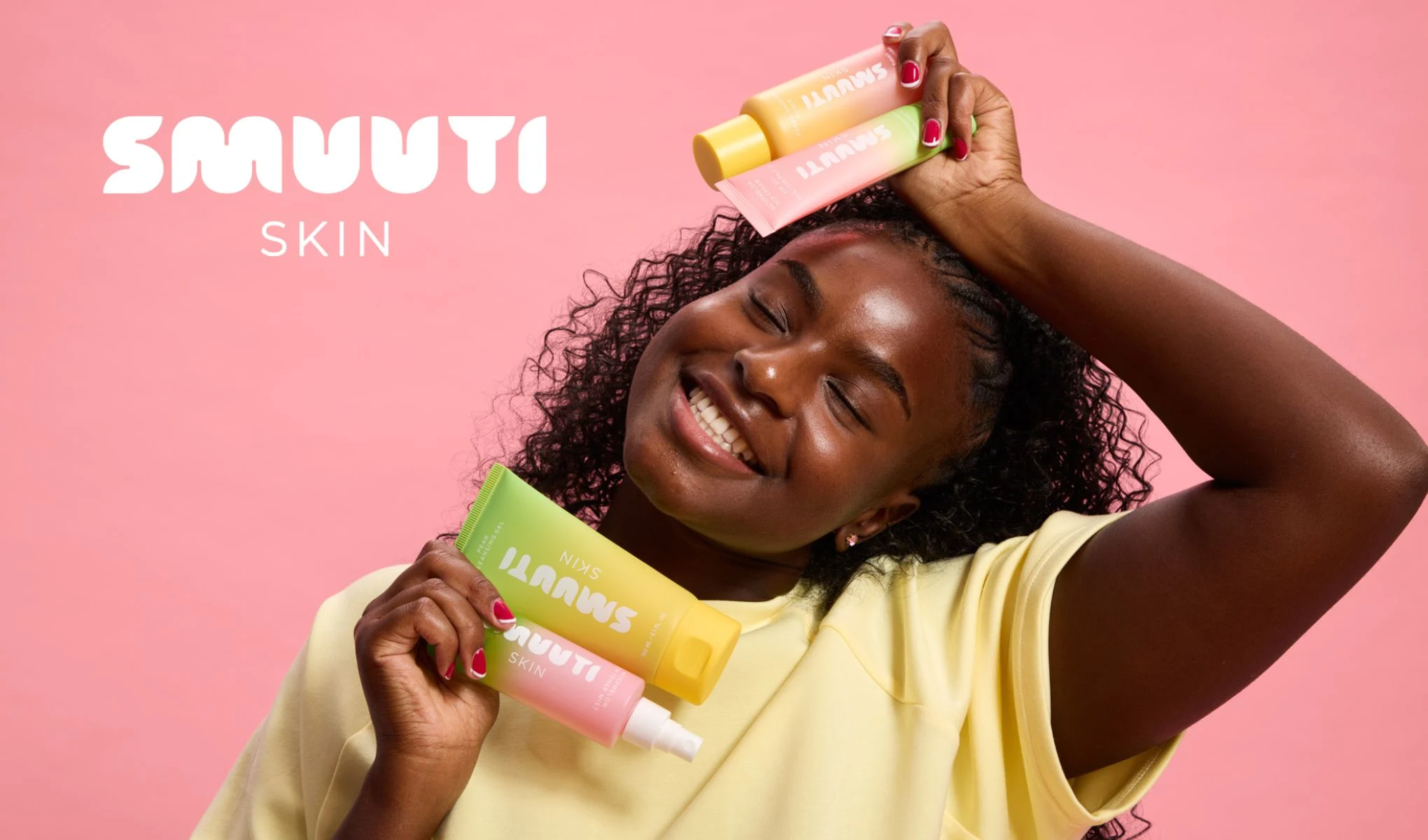 En person håller tre SMUUTI SKIN-produkter framför en rosa bakgrund. Personen har på sig en gul tröja och har röda naglar.
