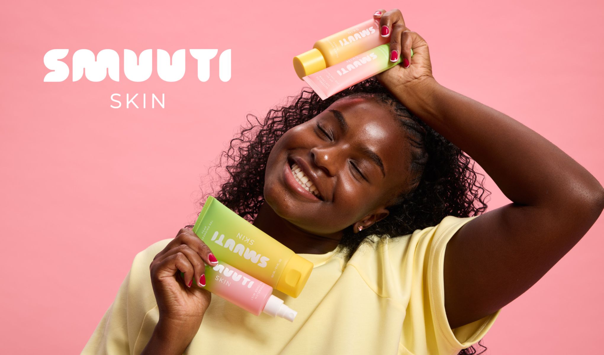 En person holder tre SMUUTI SKIN-produkter foran en rosa bakgrunn. Personen har på seg en gul skjorte og har røde negler.