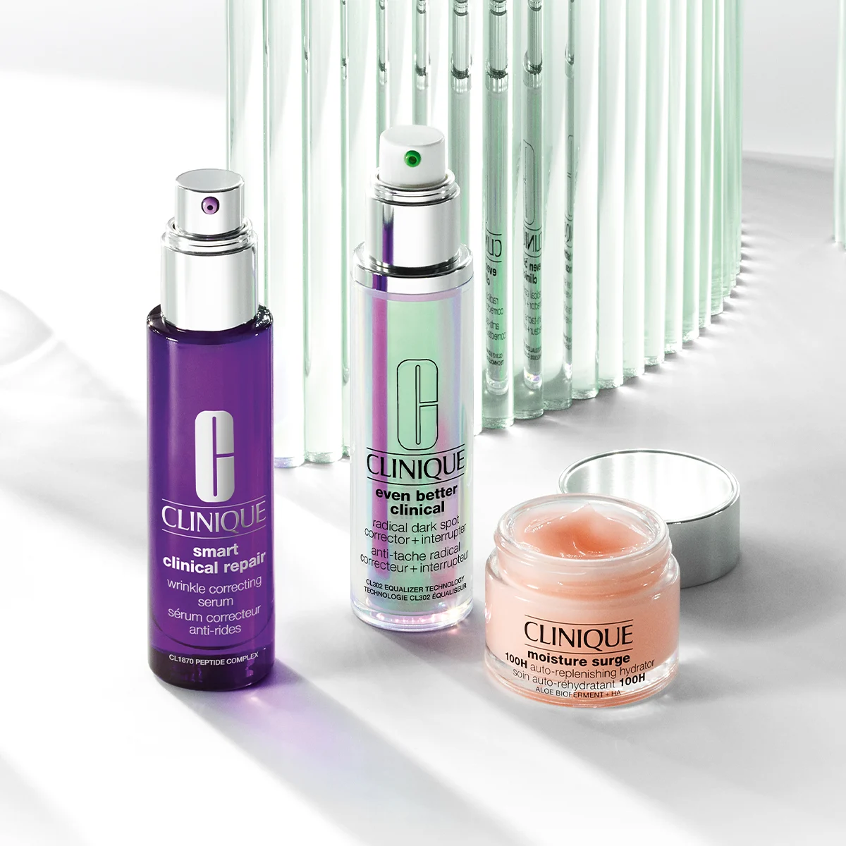 Tre hudpleieprodukter fra Clinique – to pumpeflasker og en åpen krukke – plassert på en lys overflate med refleksjoner.