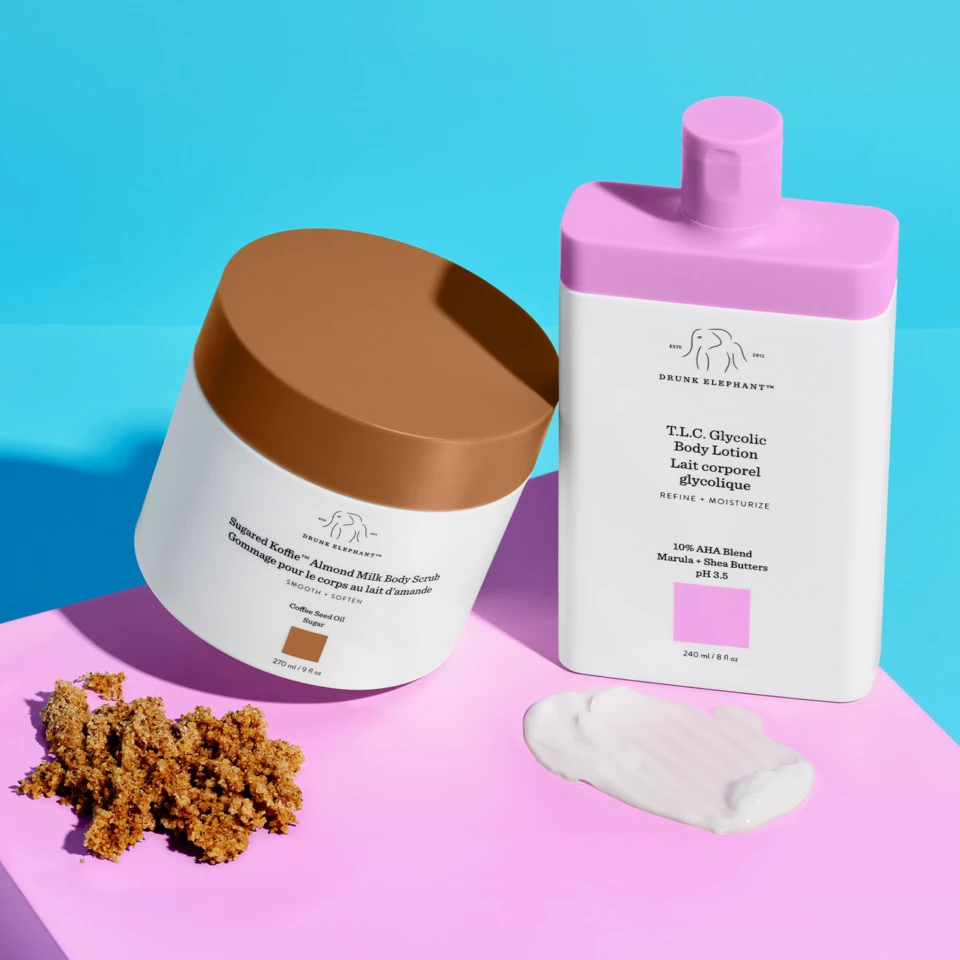 To Drunk Elephant kroppspleieprodukter: en kroppsskrubb med brunt lokk og en kroppslotion med rosa topp, plassert på en rosa og blå bakgrunn med produktteksturer ved siden av.