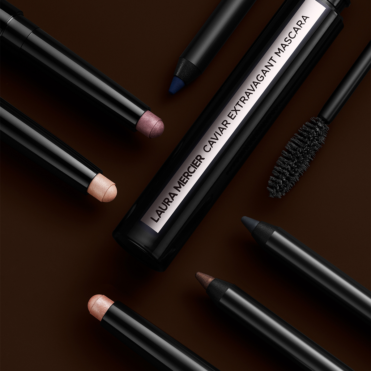 Laura Mercierin Caviar Extravagant Mascara, jota ympäröivät erilaiset silmämeikin tuotteet, mukaan lukien silmänympärys- ja silmänympärysvarjostimet lyijykynän muodossa, tummalla pohjalla.