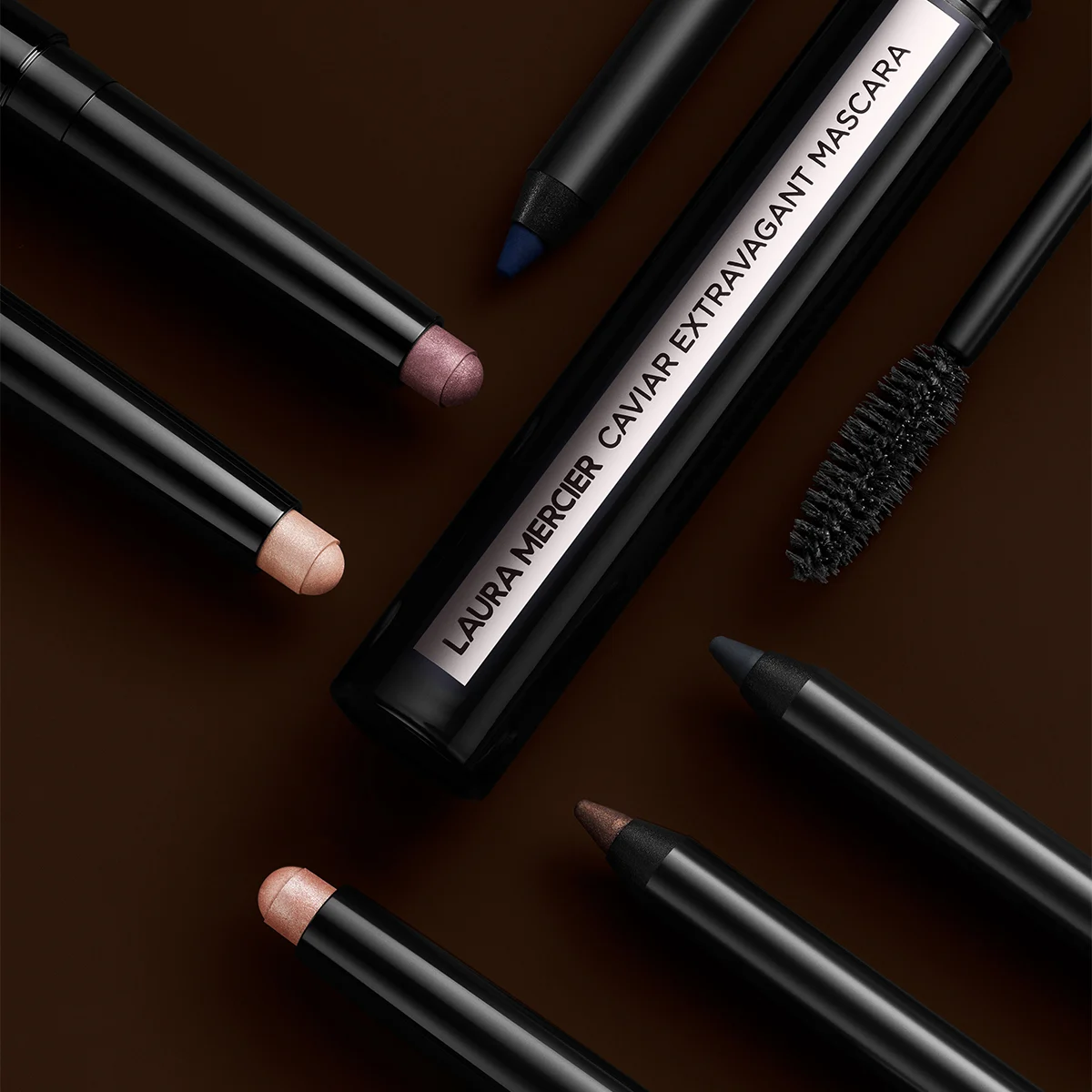 Laura Merciers Caviar Extravagant Mascara omgiven av olika ögonprodukter, inklusive ögonpennor och ögonskuggor i stiftform, på en mörk bakgrund.