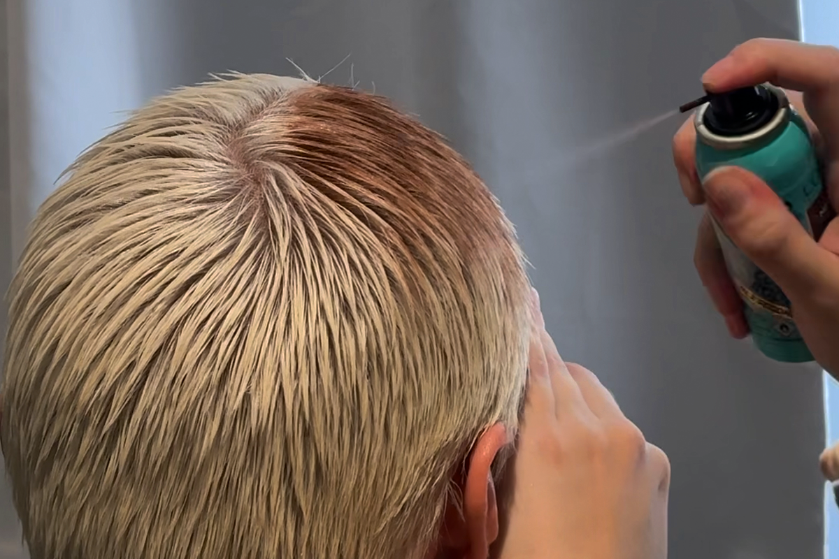 En person med blondt hår som sprayer håret med en hårfargespray.