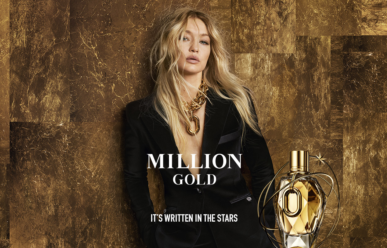 En elegant person klädd i svart kostym och guldsmycken står framför en bakgrund med guldtoner. Till höger syns en guldfärgad parfymflaska.