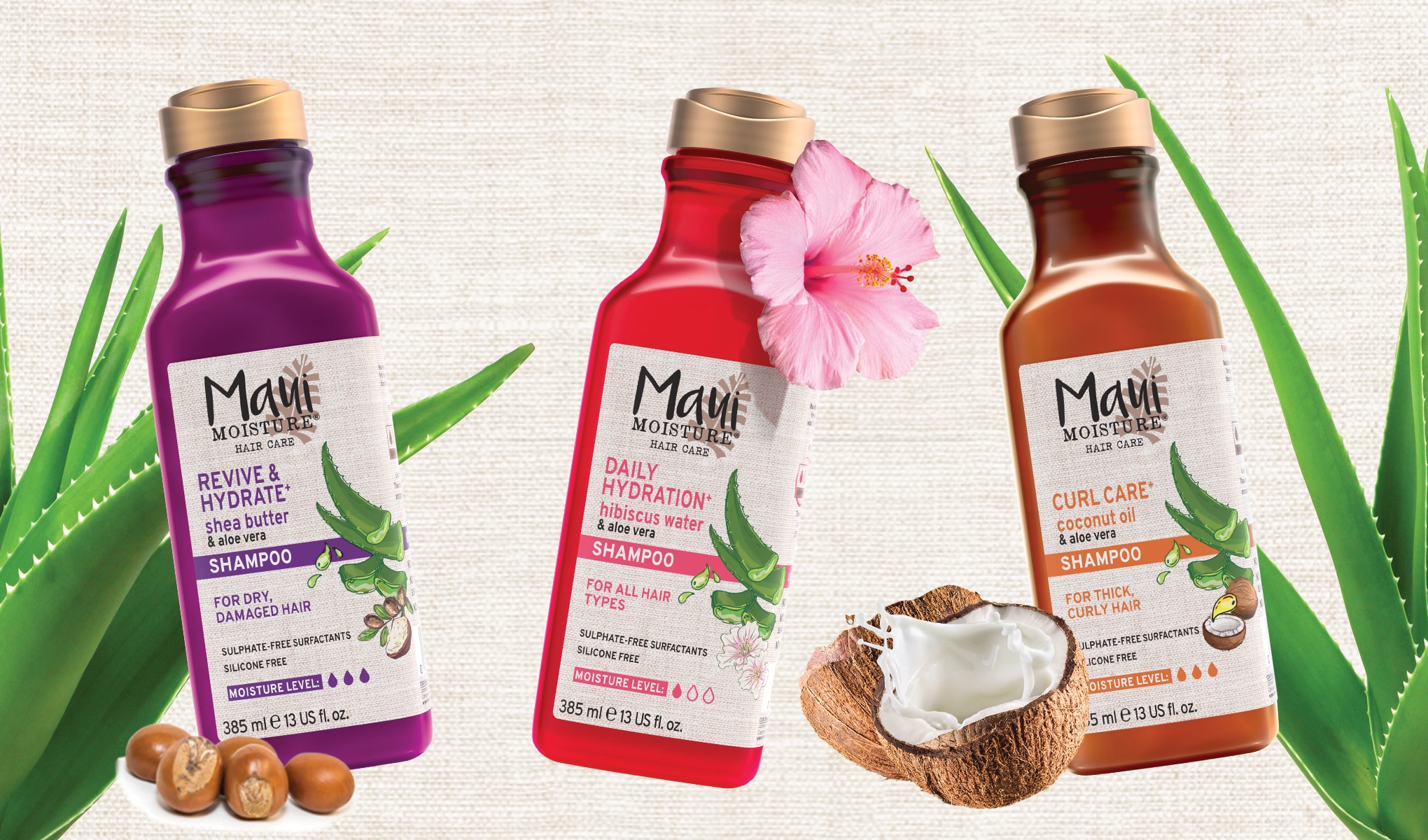 Tre Maui Moisture-schampoflaskor på ljus bakgrund: lila, röd och brun. Omringade av aloe vera, sheanötter, hibiskusblomma och kokosnöt.