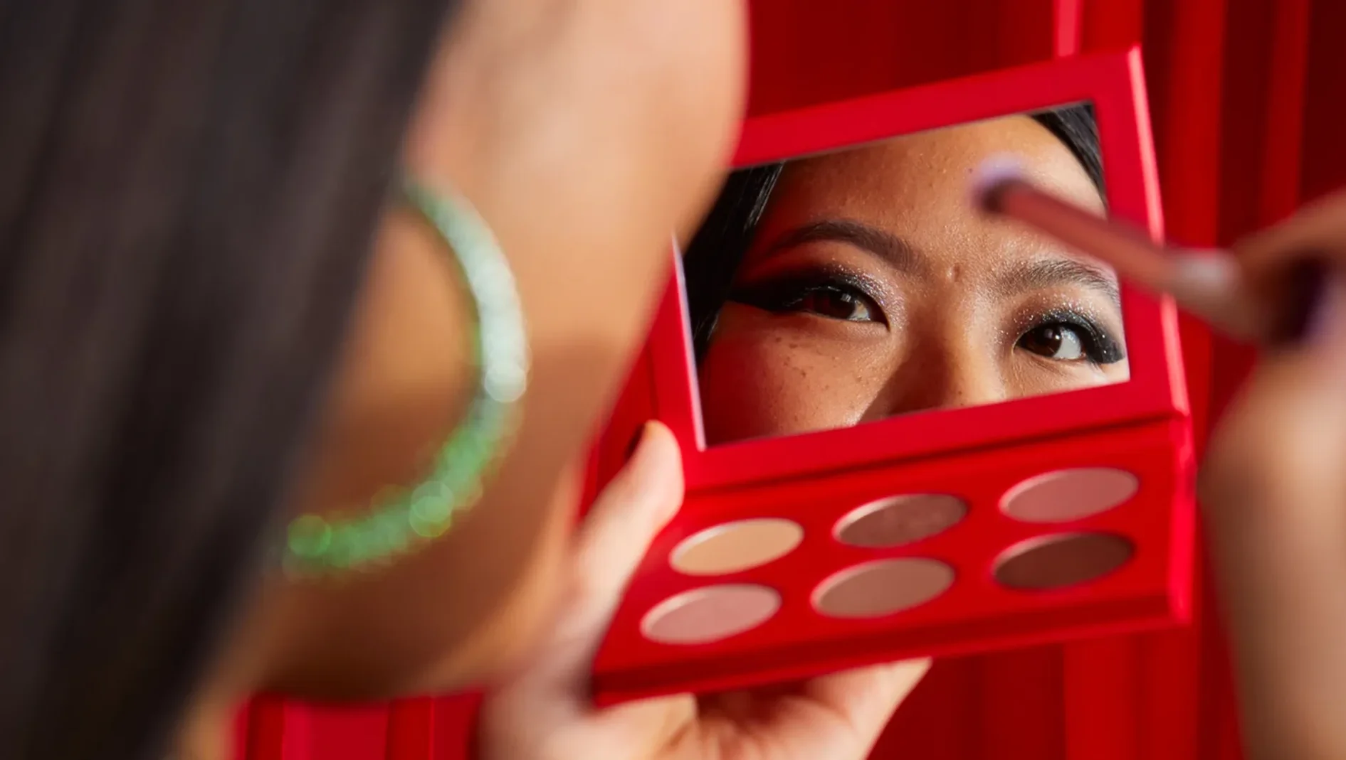 Person håller en röd ögonskuggspalett med sex nyanser och ser i palettens spegel medan ögonmakeup appliceras med en sminkborste.