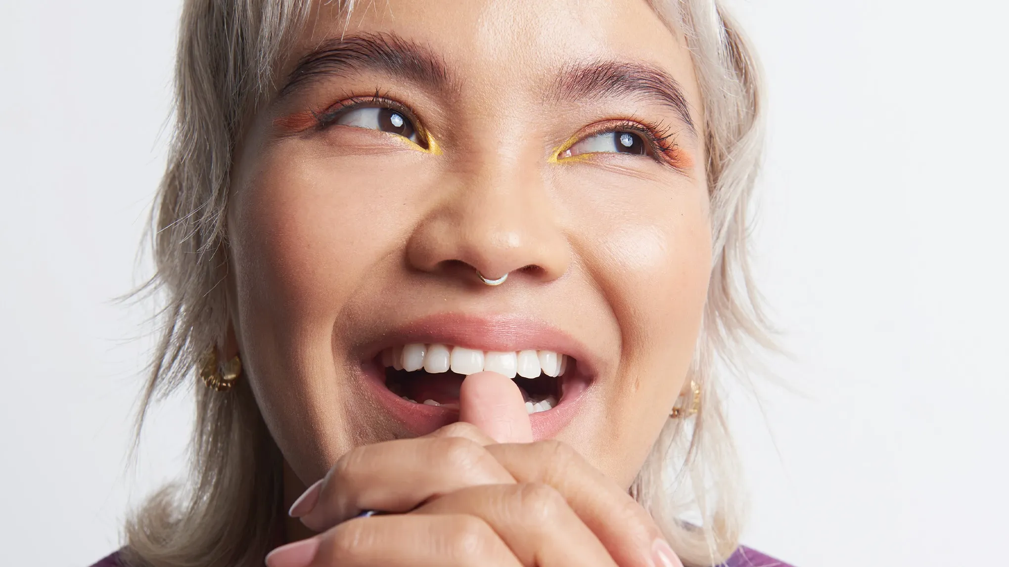 Kvinne med sminke, nesepiercing og kort, blondt hår, som smiler med åpen munn, ser til siden og har hendene knyttet foran haken mot en hvit bakgrunn.