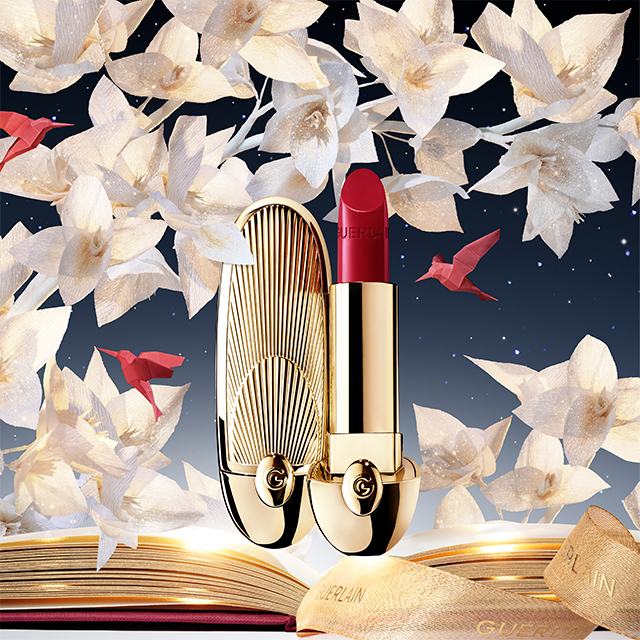 Ett elegant rött läppstift från Guerlain med en lyxig guldförpackning. Läppstiftet har en djup röd färg och förpackningen är dekorerad med en röd texturerad yta som liknar krokodilmönster. På förpackningen finns en liten "G" ingraverad. Läppstiftet är öppet och visar den vackra röda färgen, medan förpackningen ligger bredvid. Designen är sofistikerad och exklusiv.