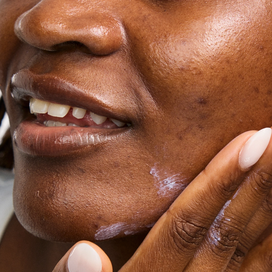 Närbild av kind där en hand applicerar hudkräm på huden med naturlig lyster.