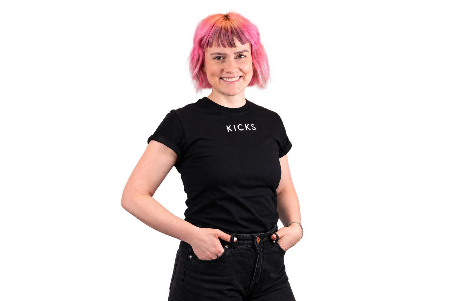 Person med kort rosa hår står foran en lys grå bakgrunn og har på seg en svart T-skjorte med hvit KICKS-logo samt mørke jeans. Portrettbilde med avslappet positur og hendene i lommene.