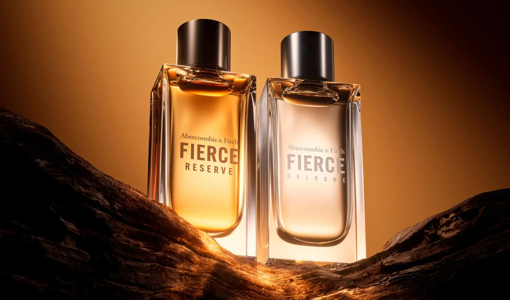 Två Abercrombie & Fitch Fierce-parfymflaskor på en träyta. Till vänster Fierce Reserve med gyllene vätska, till höger Fierce Cologne med klar vätska. Bakgrunden är orangebrun.