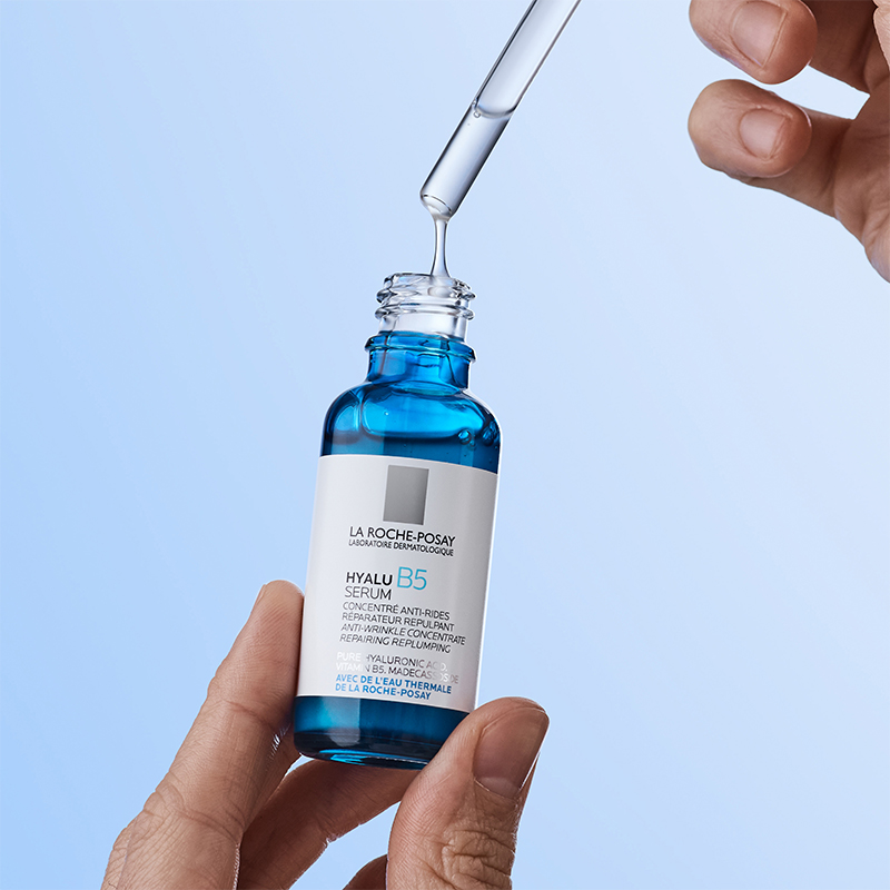 En hand håller en blå flaska med La Roche-Posay Hyalu B5 Serum, medan en annan hand använder en pipett för att droppa serumet. Bakgrunden är ljusblå och minimalistisk, vilket framhäver produktens återfuktande och reparerande egenskaper.