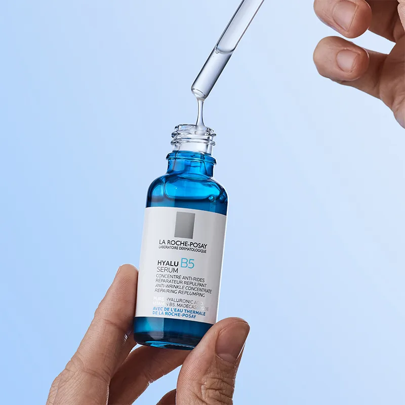 En hand håller en blå flaska med La Roche-Posay Hyalu B5 Serum, medan en annan hand använder en pipett för att droppa serumet. Bakgrunden är ljusblå och minimalistisk, vilket framhäver produktens återfuktande och reparerande egenskaper.