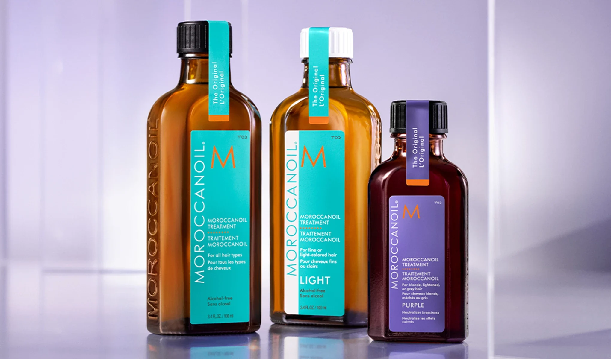 Tre flaskor med Moroccanoil-behandling står på en reflekterande yta. Flaskorna har etiketter i turkos och lila, och är märkta med "The Original" och "Alcohol-free".