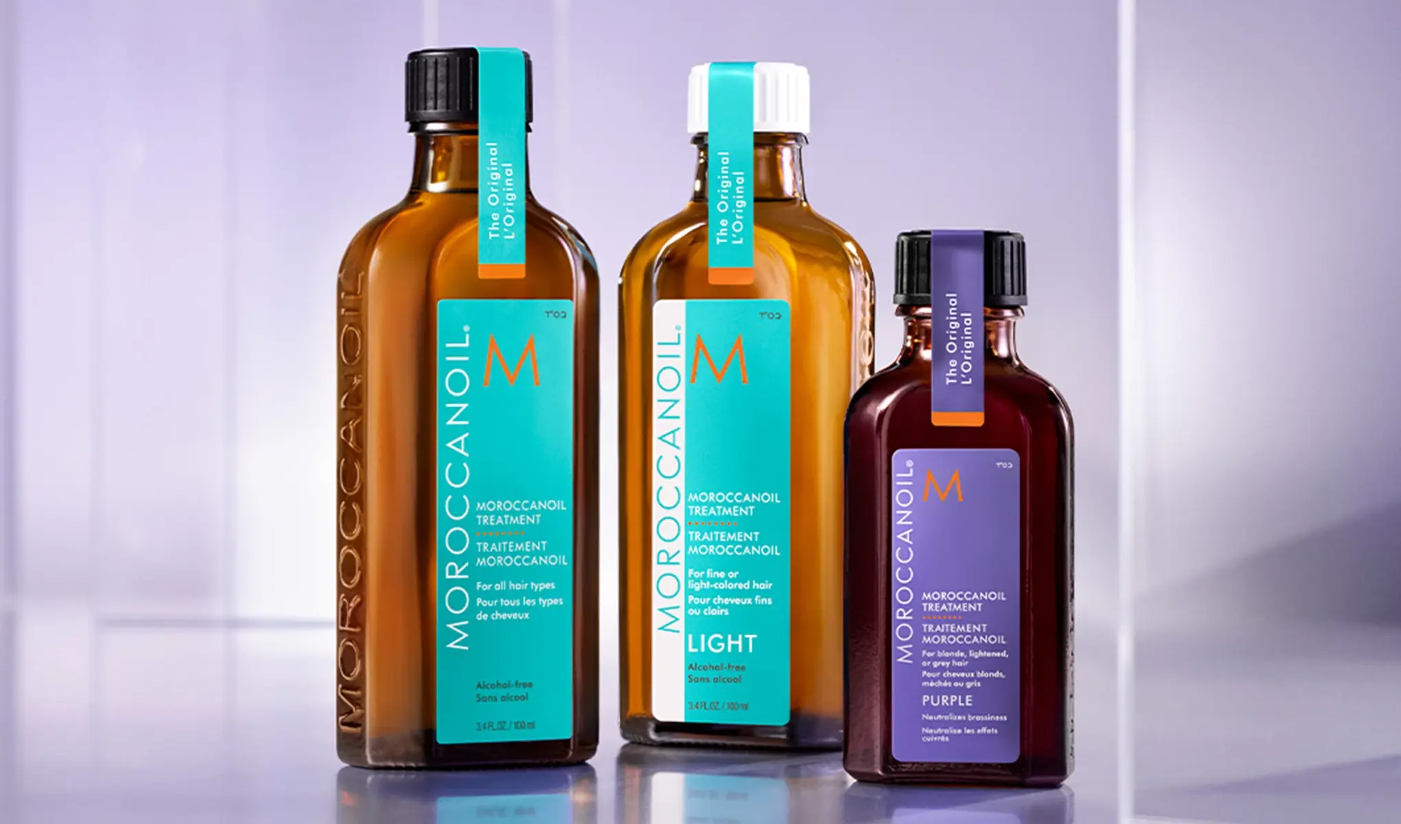 Tre flasker med Moroccanoil-behandling står på en reflekterende overflate. Flaskene har etiketter i turkis og lilla, og er merket med "The Original" og "Alcohol-free".