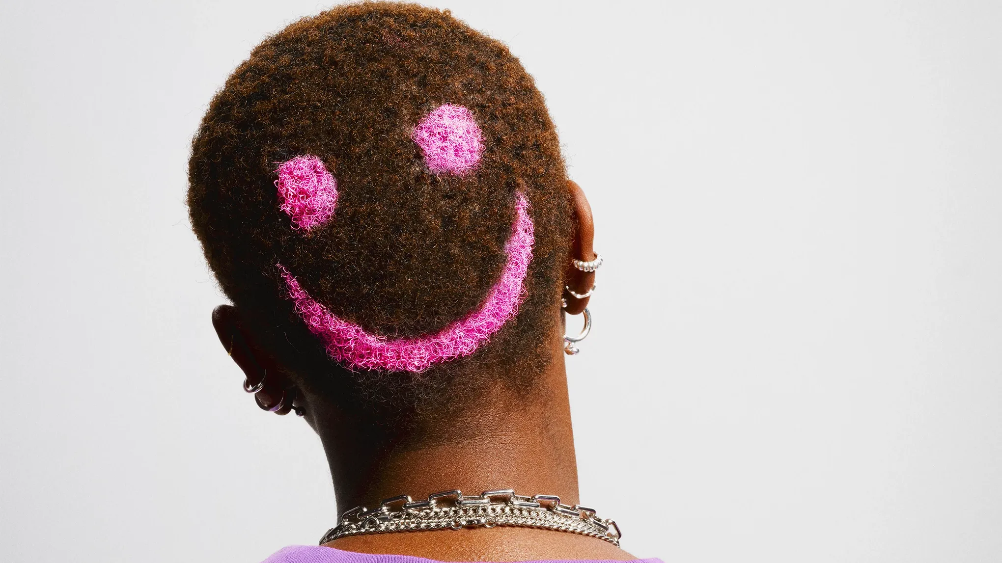 Person bakfra med snauklipt hår og hodet på skrå, mot en hvit bakgrunn. Håret er farget med en rosa smiley i bakhodet.