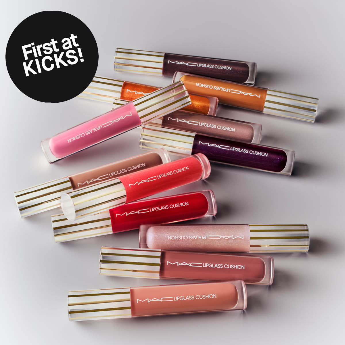 Flera läppglans från MAC Lipglass Cushion i olika nyanser ligger utplacerade mot ljus bakgrund, med guldrandiga korkar. Text på produkterna visar “MAC Lipglass Cushion” och en svart cirkel med “First at KICKS!”.